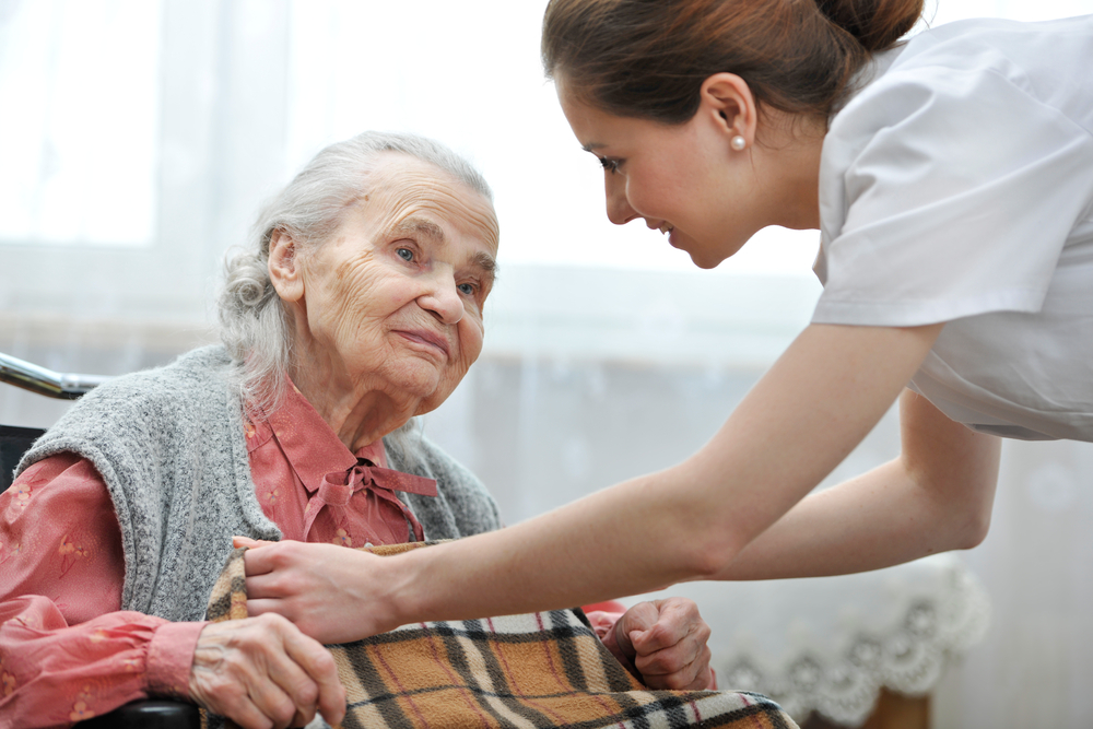 Old woman is receiving care from a nurse who is putting n her a blanket to keep her warm , they are looking at each other with gratitude